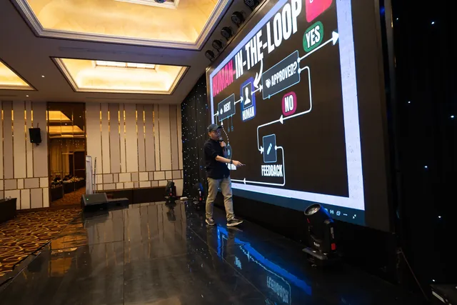 Daniel on stage beside a screen showing a human-in-the-loop approval flow