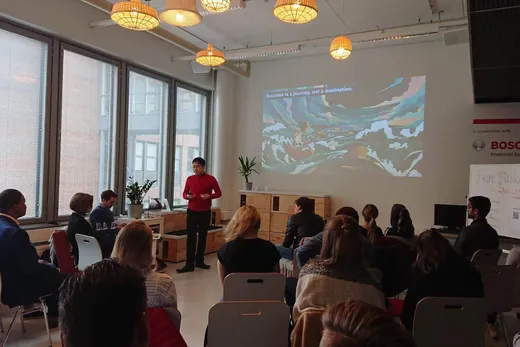 Daniel presenting to a seated audience in a bright room with large windows